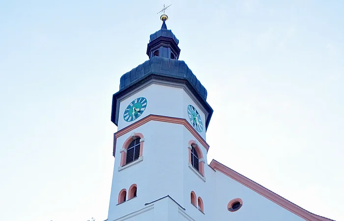 Blick durch ein Metalltor auf eine weiße Kirche mit Turmuhr und Treppe, im Hintergrund Bäume.