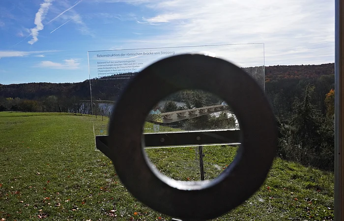 Blick durch ein rundes Metallrohr auf eine Infotafel mit Brückenrekonstruktion vor einer Wiese und Wald im Hintergrund.