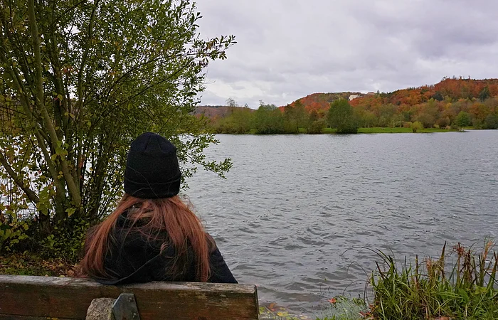 Person mit schwarzer Mütze sitzt auf Bank und blickt auf See mit herbstlicher Landschaft im Hintergrund.
