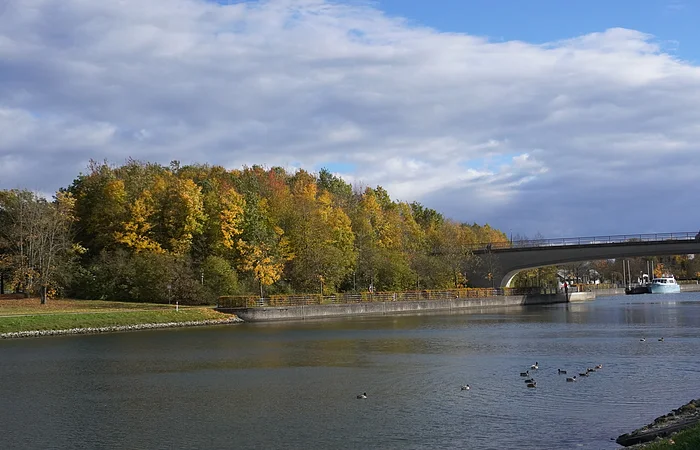 Fluss mit Enten, Brücke und herbstlich gefärbten Bäumen unter bewölktem Himmel.