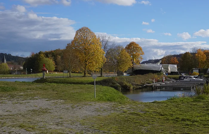 Uferbereich mit Bootssteg, herbstlich gefärbten Bäumen und bewölktem Himmel im Hintergrund.