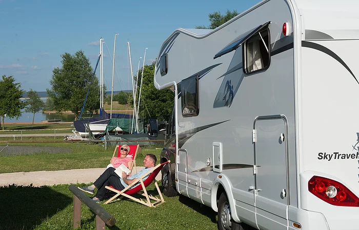 Eine Frau und ein Mann sitzen in zwei Liegestützen link neben einem Wohnmobil. Im Hintergund lagern Boote auf Anhängern. Dahinter beginnt der See.