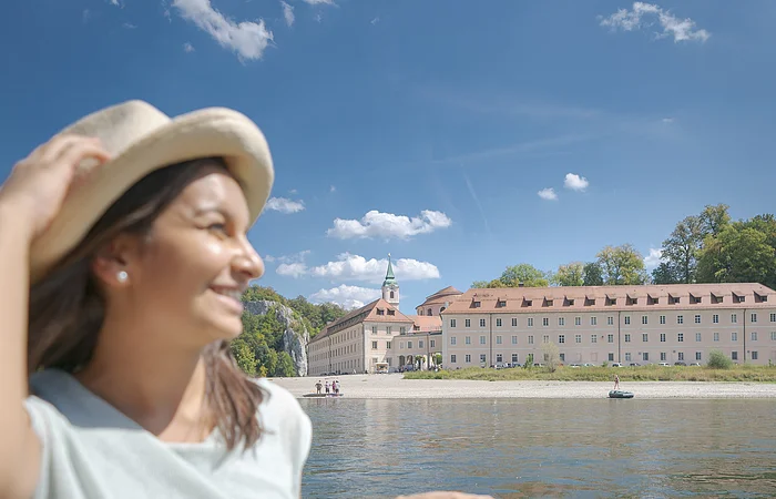 Eine junge Frau mit Strohhut und sommerlicher Kleidung sitzt in einem Ausflugsschiff mit grüner Sitzbank. Neben ihr liegt die Sonnenbrille auf der Bank. Im Hintergrund ist die Donau und das Kloster Weltenburg zu sehen. Am hellblauen Himmel sind einige weiße Schäfchenwolken zu sehen.