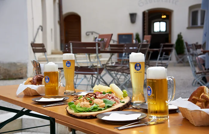 Weizengläser und Halbekrüge mit Bier. Brotzeitplatte mit Bretzel, Käse und Wurst auf Holztisch im Biergarten vor Stühlen und Gebäudeeingang.