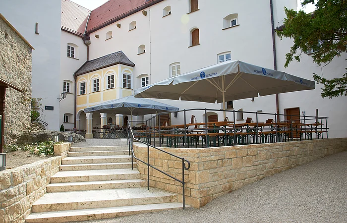 Außenbereich mit Treppe, Sitzbänken und Sonnenschirmen vor einem historischen Gebäude.