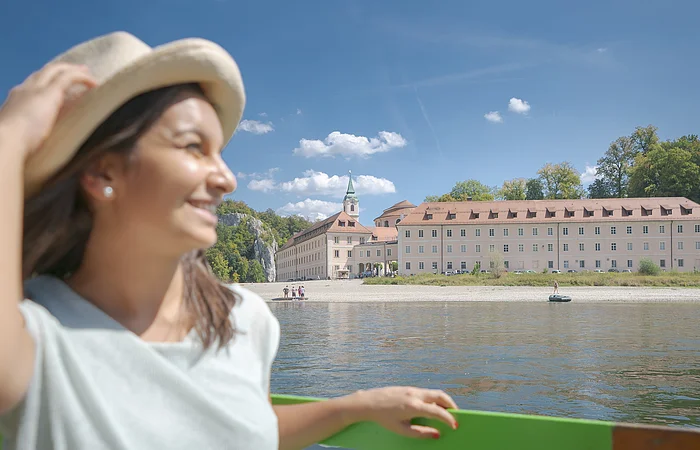 Eine junge Frau mit Strohhut und sommerlicher Kleidung sitzt in einem Ausflugsschiff mit grüner Sitzbank. Neben ihr liegt die Sonnenbrille auf der Bank. Im Hintergrund ist die Donau und das Kloster Weltenburg zu sehen. Am hellblauen Himmel sind einige weiße Schäfchenwolken zu sehen.