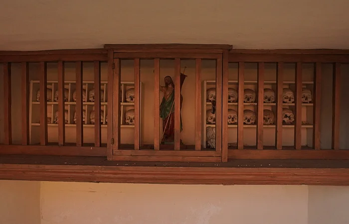 Holzregal mit mehreren Totenschädeln und einer Heiligenfigur dahinter hinter Holzgittern in einer Wandnische.