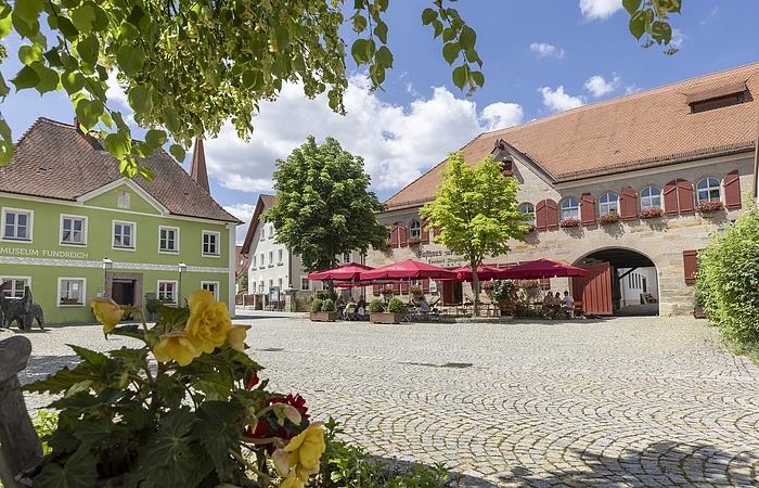 Pflanzen im Vordergrund, gepflasterter Platz mit Bäumen und Sitzbereich vor historischen Gebäuden bei Sonnenschein.