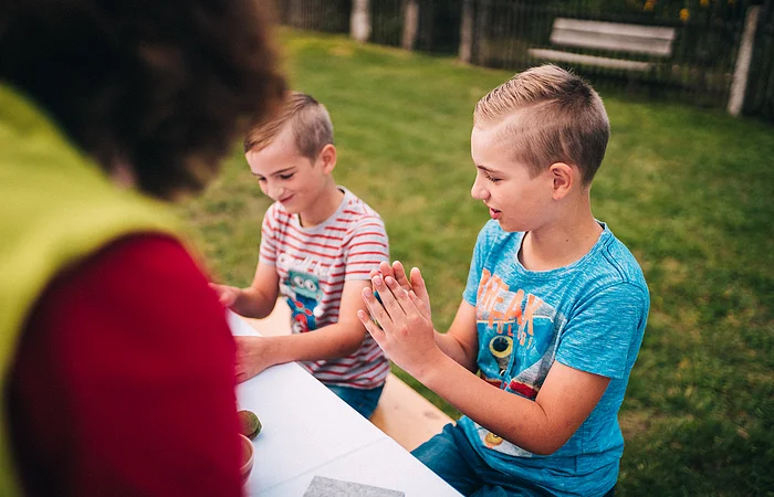 Zwei Jungen sitzen an einem Tisch im Freien, ein Erwachsener ist unscharf im Vordergrund zu sehen.