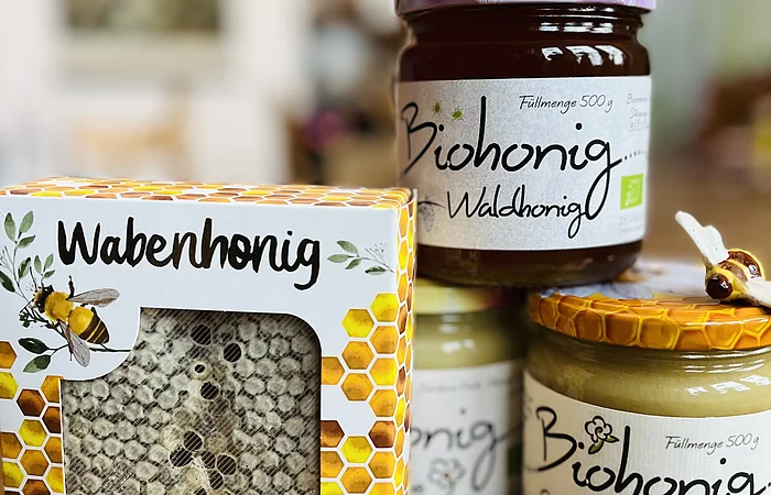 Vier Honigprodukte, darunter Wabenhonig in Verpackung und drei Gläser Biohonig, auf Holztisch.