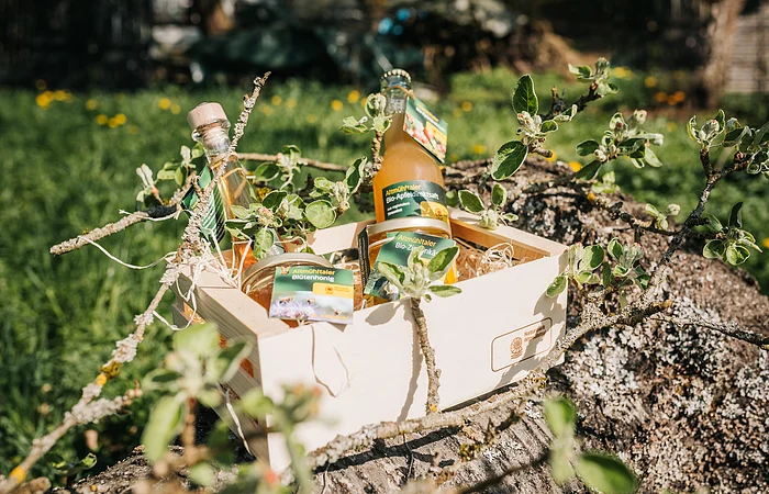 Holzkiste mit Bio-Produkten, darunter Apfelsaft, Honig und Likör, auf einem Baumstamm im Freien platziert.