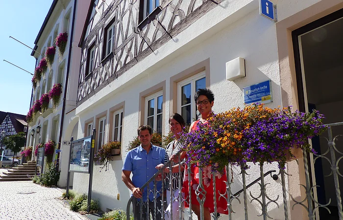 Drei Personen stehen auf einer Treppe vor einem Gebäude, der Touristinformation Monheim. Das Gebäude mit Fachwerkfassade und Blumenkästen steht auf einem gepflasterten Platz, daneben ein blaues Schild mit der Aufschrift "Tourist-Info". Im Hintergrund ein weiteren dreistöckiges Gebäude mit Blumenkästen.