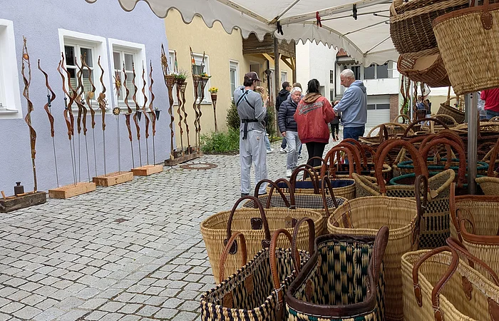Marktstand in einer Innenstadt mit geflochtenen Körben, im Hintergrund Menschen und dekorative Metallstäbe vor bunten Häuserwänden.