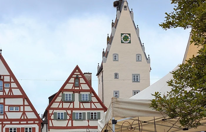 Markt mit Ständen und Besuchern vor Fachwerkhäusern und einem Turm mit Uhr und Storchnest.