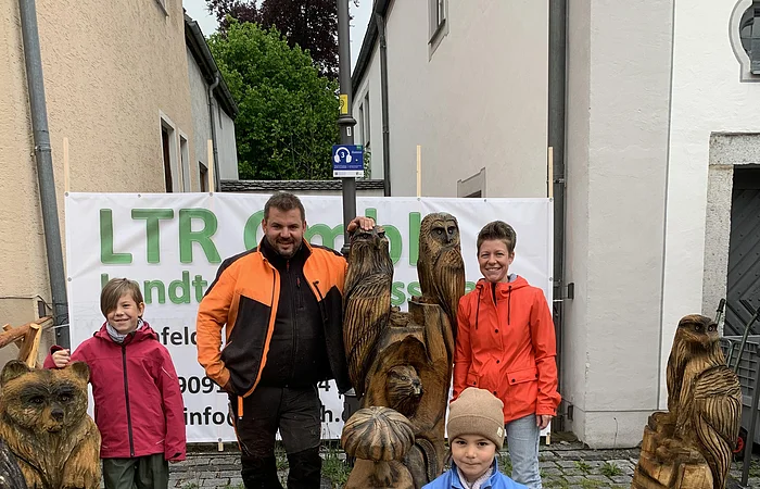 Vier Personen stehen hinter und neben geschnitzten Holzfiguren von Tieren auf einem gepflasterten Platz und blicken in die Kamera. Im Hintergrund zwei Wohnhäuser und eine kleine Gasse zu sehen, sowie ein Werbebanner an einem Bauzaun.