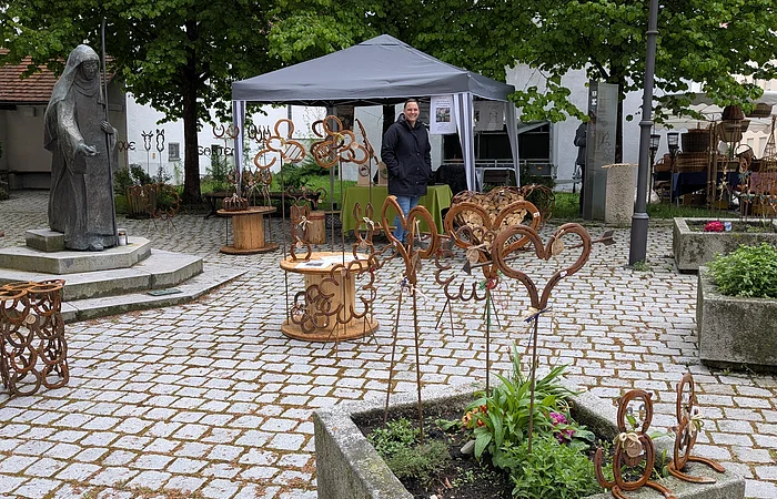 Marktstand mit Metallkunstwerken auf dem Pflasterplatz verteilt, Person am Pavillon mit schwarzem Dach, Marienstatue links, Bäume und Wohnhäuser im Hintergrund.