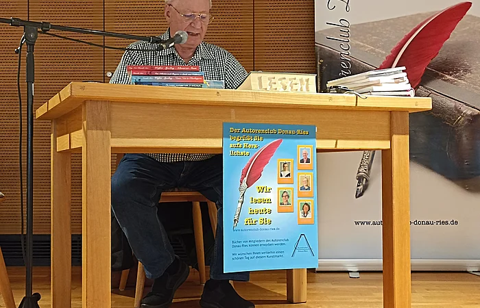 Älterer Mann liest an einem Holztisch mit Mikrofon, Plakat des Autorenclubs Donau-Ries hängt am Tisch. Neben dem Tisch ein großer Aufsteller des Autorenclubs Donau-Ries.