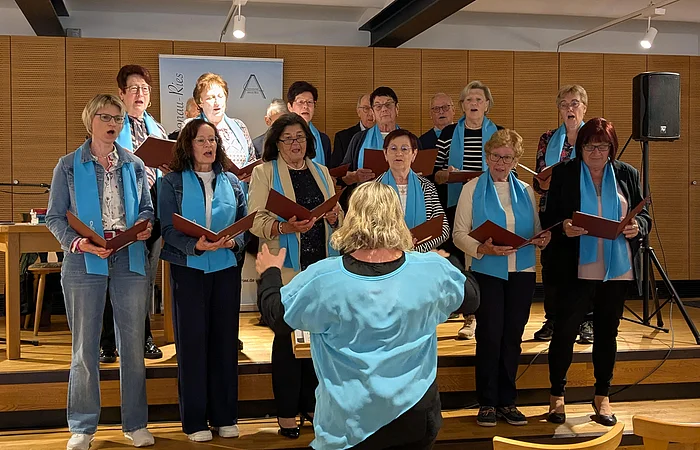 Chor mit 15 Personen und Dirigentin, alle mit blauen Schals um den Hals, singen auf einer Bühne in einem Raum mit Holzverkleidung im Hintergrund. Vor ihnen stehen Holzstühle.