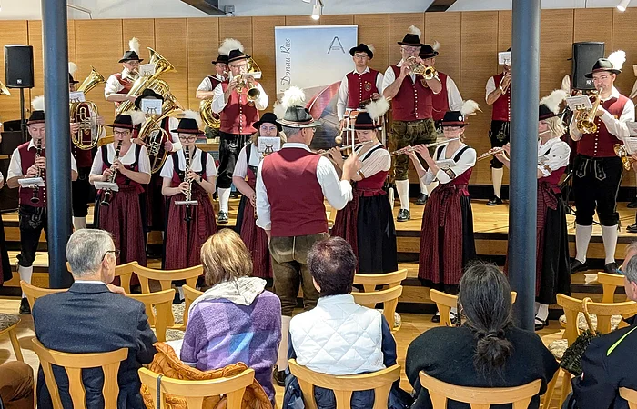 Blaskapelle in rot/schwarz/weißer Tracht spielt auf der Bühne, mit einer Holzverkleidung im Hintergrund, davor sitzen sechs Zuschauer auf Holzstühlen.