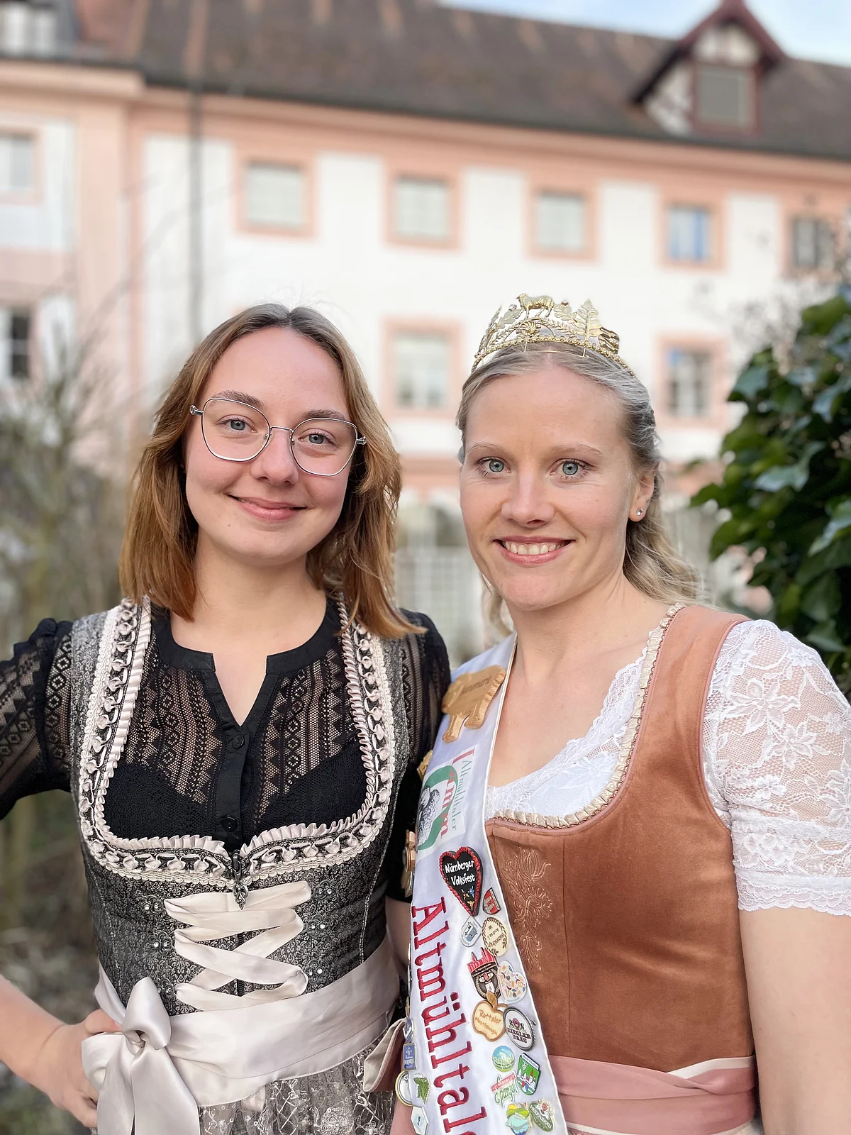 Amtsübergabe bei der Altmühltaler Lammkönigin Zwei Frauen in traditionellen Trachten, eine mit Krone und Schärpe, vor einem Gebäude mit Fenstern.