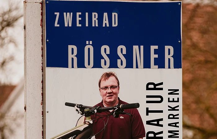 Werbeschild für Fahrradreparatur mit Mann und Fahrrad, Text „Zweirad Rössner Reparatur aller Marken“ und Telefonnummer.