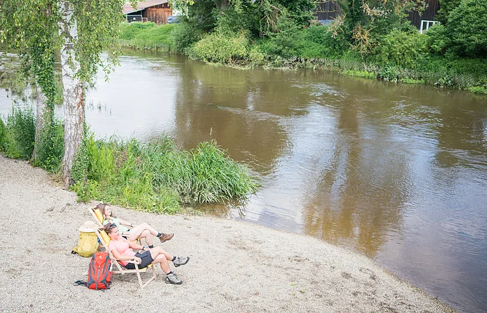 Zwei Personen mit Rucksäcken neben sich  sitzen auf Liegestühlen am Flussufer neben Bäumen an einem sonnigen Tag.