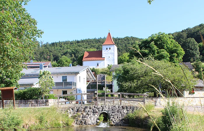 Fluss mit klarstem Wasser, Brücke aus Stein, Häuser und Kirche mit rotem Dach vor bewaldetem Hügel unter blauem Himmel.