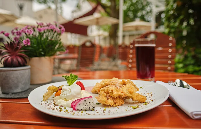Auf einem Holztisch auf der Terrasse ist eine köstlich angerichtete Portion Obatzda serviert. Im Hintergrund stehen links zwei kleine Blumentöpfe und rechts ein Getränk.