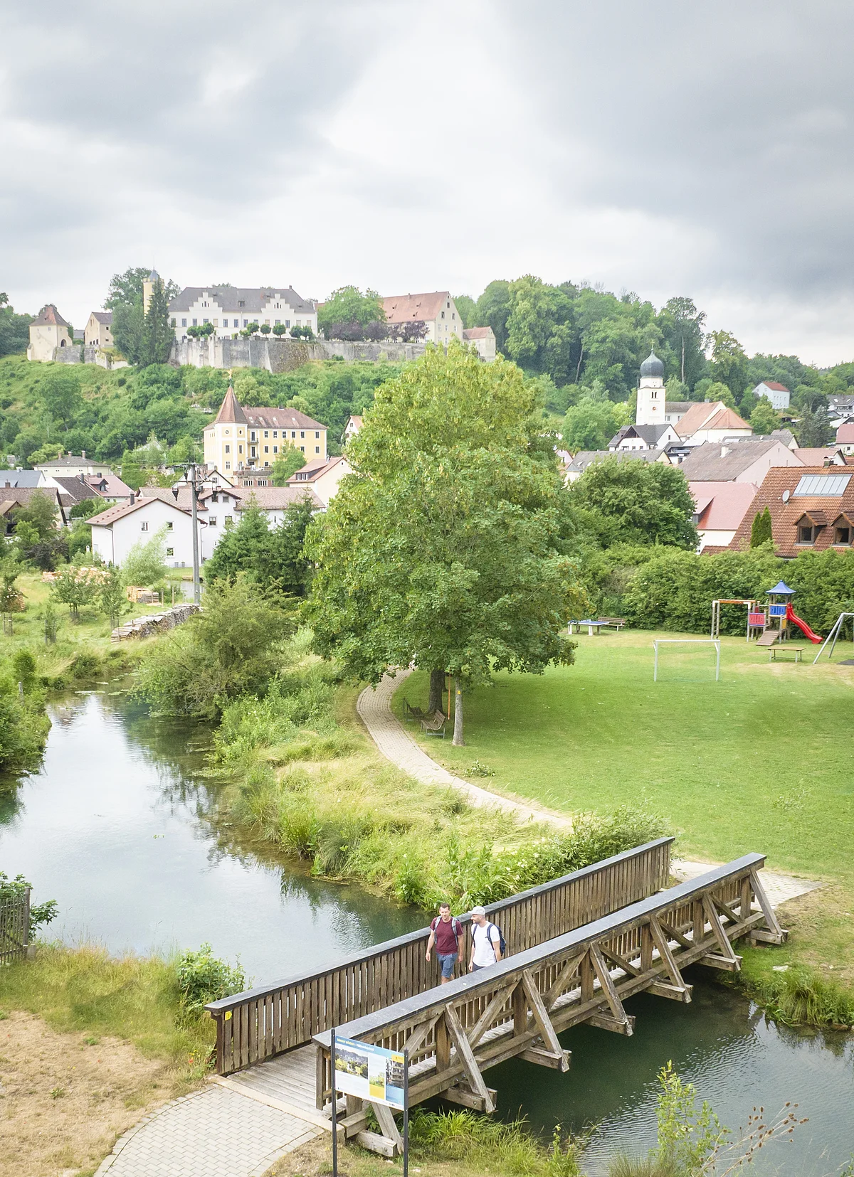 Drohnenaufnahme von Möhren. Im Vordergrund sind 2 Wanderer auf einer Holzbrücke über den Möhrenbach zu sehen. Im Hintergrund thront Schloss Möhren über dem Dorf.