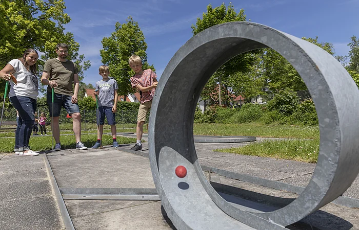 Vier Personen spielen Minigolf auf einem sonnigen Außenplatz mit Betonhindernissen und grüner Umgebung.