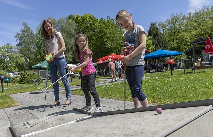Drei Personen spielen Minigolf auf einem sonnigen Platz mit Bäumen und Sonnenschirmen im Hintergrund.