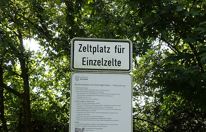 Schild mit der Aufschrift „Zeltplatz für Einzelzelte“ und weiteren Informationen vor grünem Laub.