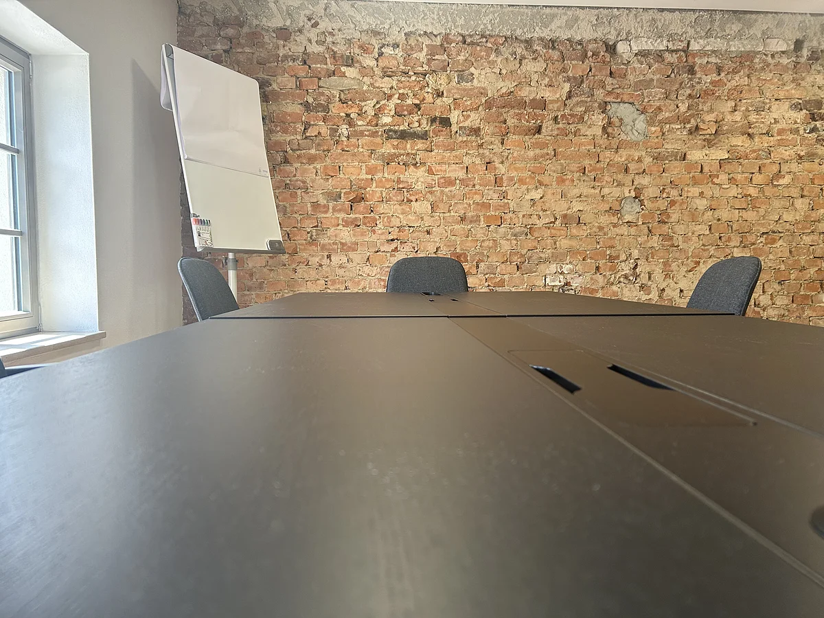 Konferenztisch mit Stühlen vor roher Backsteinwand und Flipchart neben Fenster in hellem Raum.