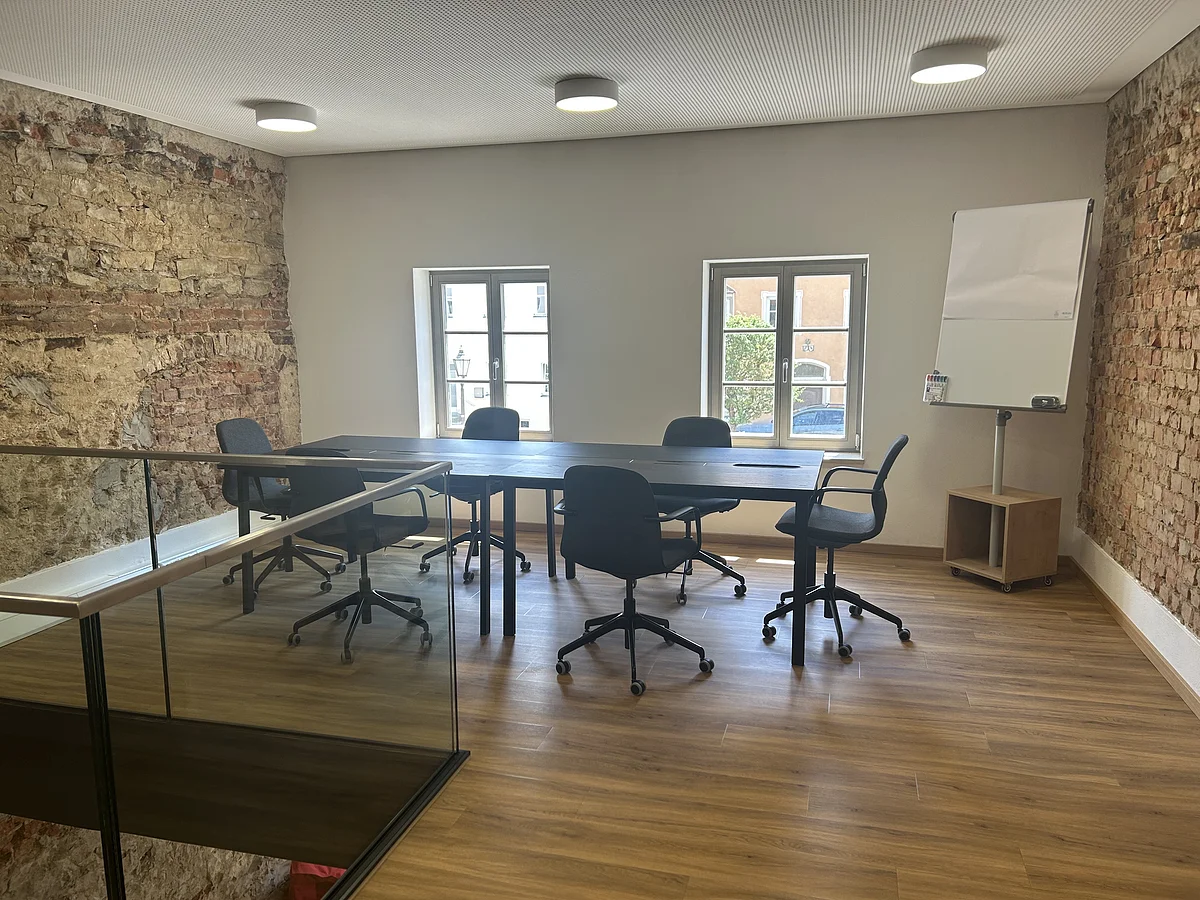 Konferenzraum mit Holzboden, sechs schwarzen Bürostühlen um einen langen Tisch, zwei Fenstern und Flipchart.