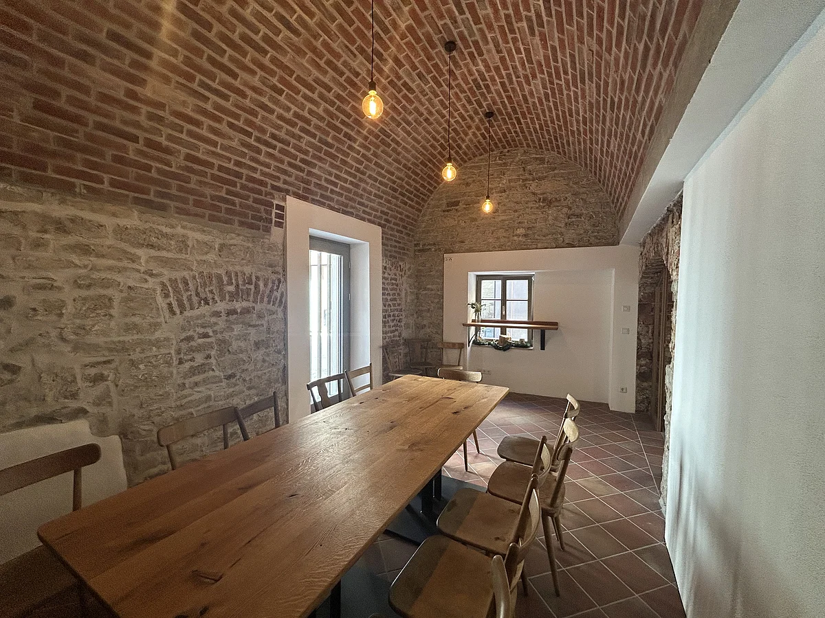 Raum mit gemauertem Tonnengewölbe, Holztisch, Holzstühlen und Fenster mit Regal und Pflanzen.