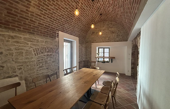Raum mit gemauertem Tonnengewölbe, Holztisch, Holzstühlen und Fenster mit Regal und Pflanzen.