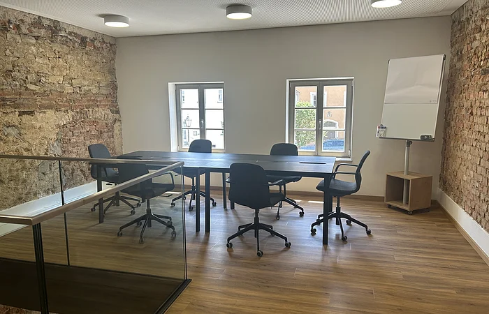 Konferenzraum mit Holzboden, sechs schwarzen Bürostühlen um einen langen Tisch, zwei Fenstern und Flipchart.