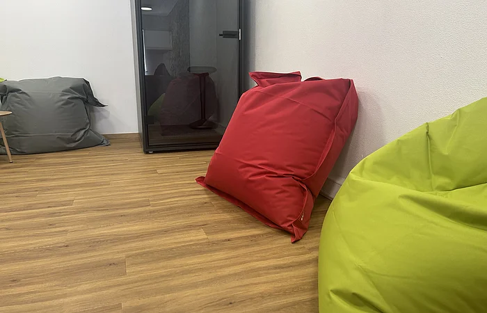 Raum mit Holzboden, weißen Wänden, grüner, roter und grauer Sitzsack und schwarzer Glasbox