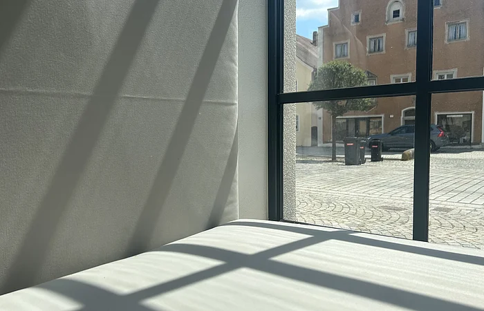 Fenster mit schwarzen Rahmen, durch das Sonnenlicht Schatten auf eine weiße Wand und Fläche wirft, draußen Gebäude und Pflastersteinweg sichtbar.