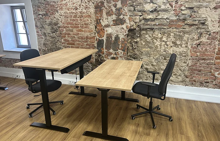 Zwei höhenverstellbare Schreibtische mit schwarzen Bürostühlen vor einer freigelegten Backsteinwand in einem Raum mit Holzboden.