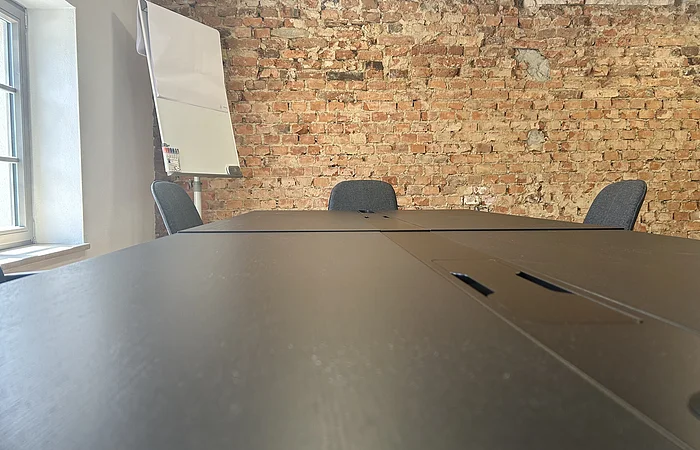 Konferenztisch mit Stühlen vor roher Backsteinwand und Flipchart neben Fenster in hellem Raum.