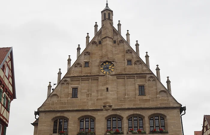 Fassade eines historischen Steingebäudes mit Uhr, Fensterbänken mit Blumen und geschlossenen Sonnenschirmen davor.