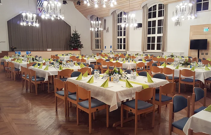 Großer Saal mit mehreren eingedeckten Tischen, Holzstühlen, Kronleuchtern und Weihnachtsbaum am Bühnenrand.