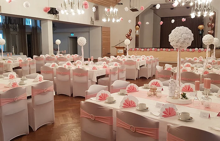 Festlich dekorierter Saal mit weißen Stuhlbezügen, rosa Schleifen, Tischen mit rosa Servietten und Kerzenständern.