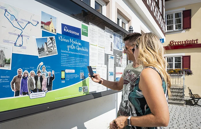 Zwei Personen vor einem weißen Gebäude, der Touristinformation Monheim betrachten eine Informationstafel mit einer Karte und Texten zur Lauschtour in Monheim und haben ein Smartphone in der Hand, mit dem sie einen Code abscannen.