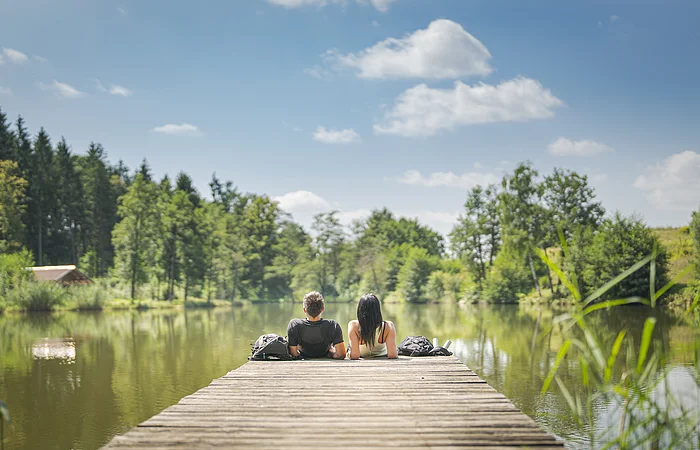 Zwei Personen sitzen auf einem Steg an einem See, umgeben von Bäumen und unter blauem Himmel. Im Hintergrund links im Bild eine kleine Holzhütte zu sehen.