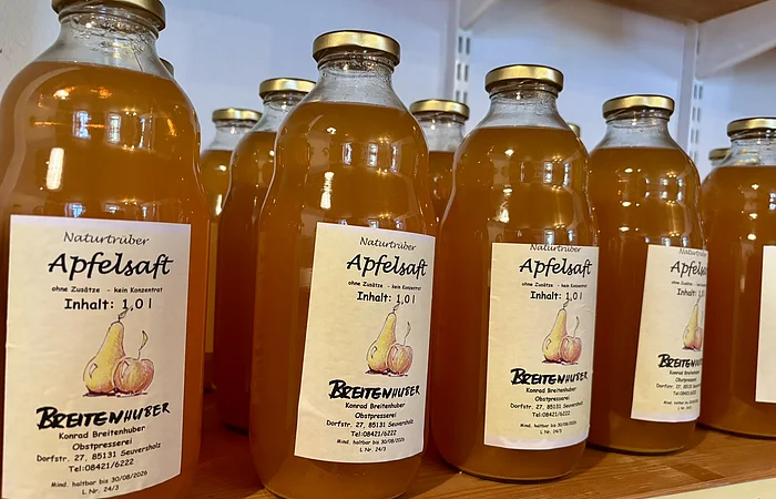 Mehrere Flaschen naturtrüber Apfelsaft mit goldfarbenen Deckeln auf einem Holzregal.