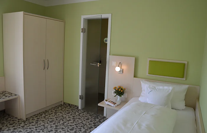Hotelzimmer mit Einzelbett, grün gestrichener Wand, Schrank, Glastür und Nachttisch mit Lampe und Blumen.