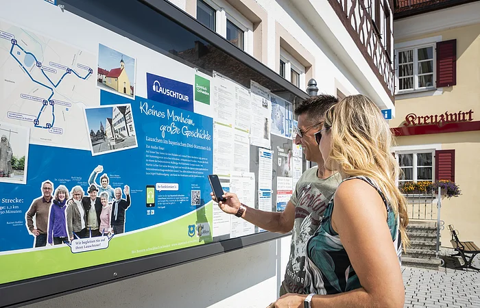 Zwei Personen vor einem weißen Gebäude, der Touristinformation Monheim betrachten eine Informationstafel mit einer Karte und Texten zur Lauschtour in Monheim und haben ein Smartphone in der Hand, mit dem sie einen Code abscannen.