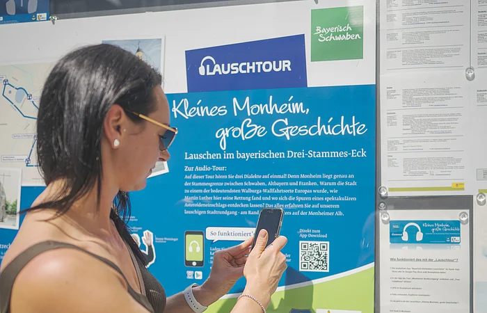 Frau mit Smartphone steht vor Infotafel zur Lauschtour in Monheim, Bayern, mit dem Titel "Kleines Monheim, große Geschichte" bei Sonnenschein. Frau hält ihr Smartphone an einen QR-Code.